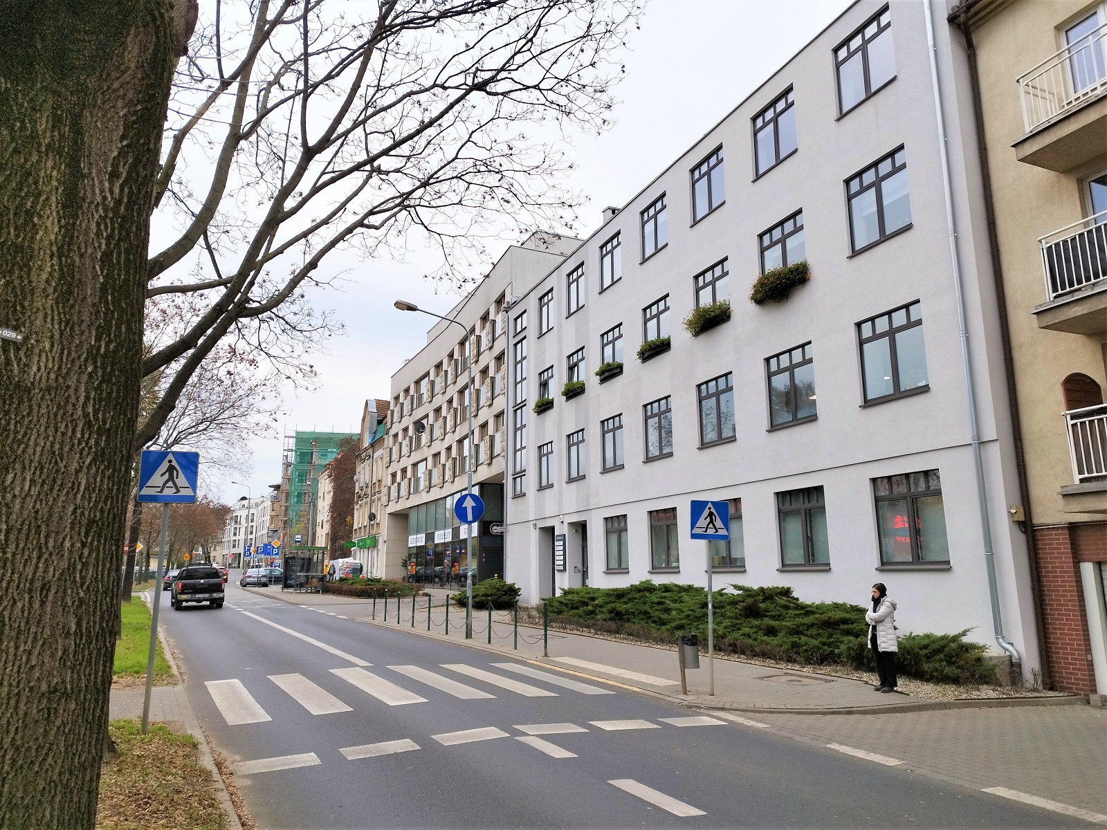 Fotografia przedstawia w centralnej części budynek w sąsiedztwie terenów zieleni urządzonej, w którym na 2 piętrze zlokalizowane jest biuro Stowarzyszenia Metropolia Poznań. Na pierwszym planie znajduje się przejście dla pieszych stosownie oznakowane dla osób z niepełnosprawnością wzroku, natomiast w tle widoczny jest przystanek autobusowy „Jeżycka” linii 164.